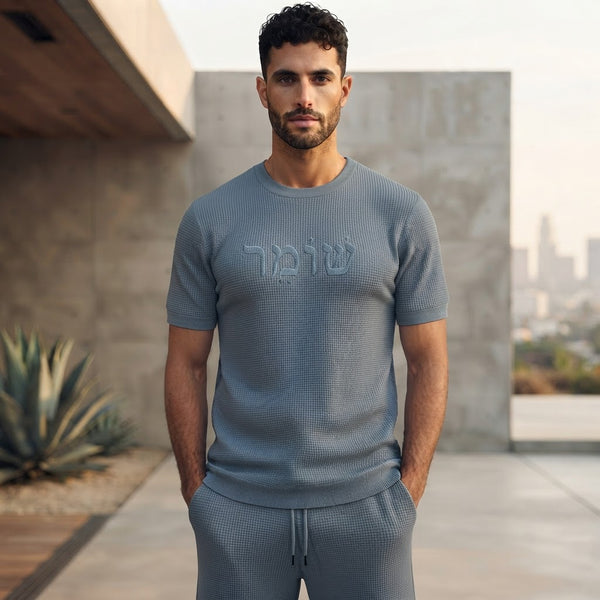 Male model wearing the Protectors men’s Shomer textured shirt and shorts set in Slate Blue, featuring tonal Hebrew שומר lettering on the chest and matching drawstring shorts.