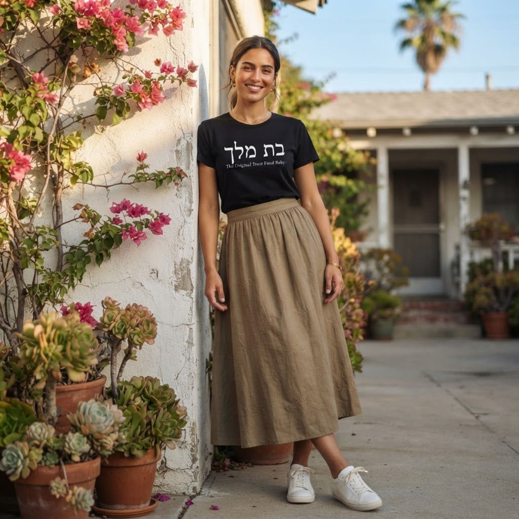 Woman wearing The Original Tee in black by Hashem Is My Sugar Daddy, featuring בת מלך and The Original Trust Fund Baby graphic styled with a long neutral skirt and white sneakers.