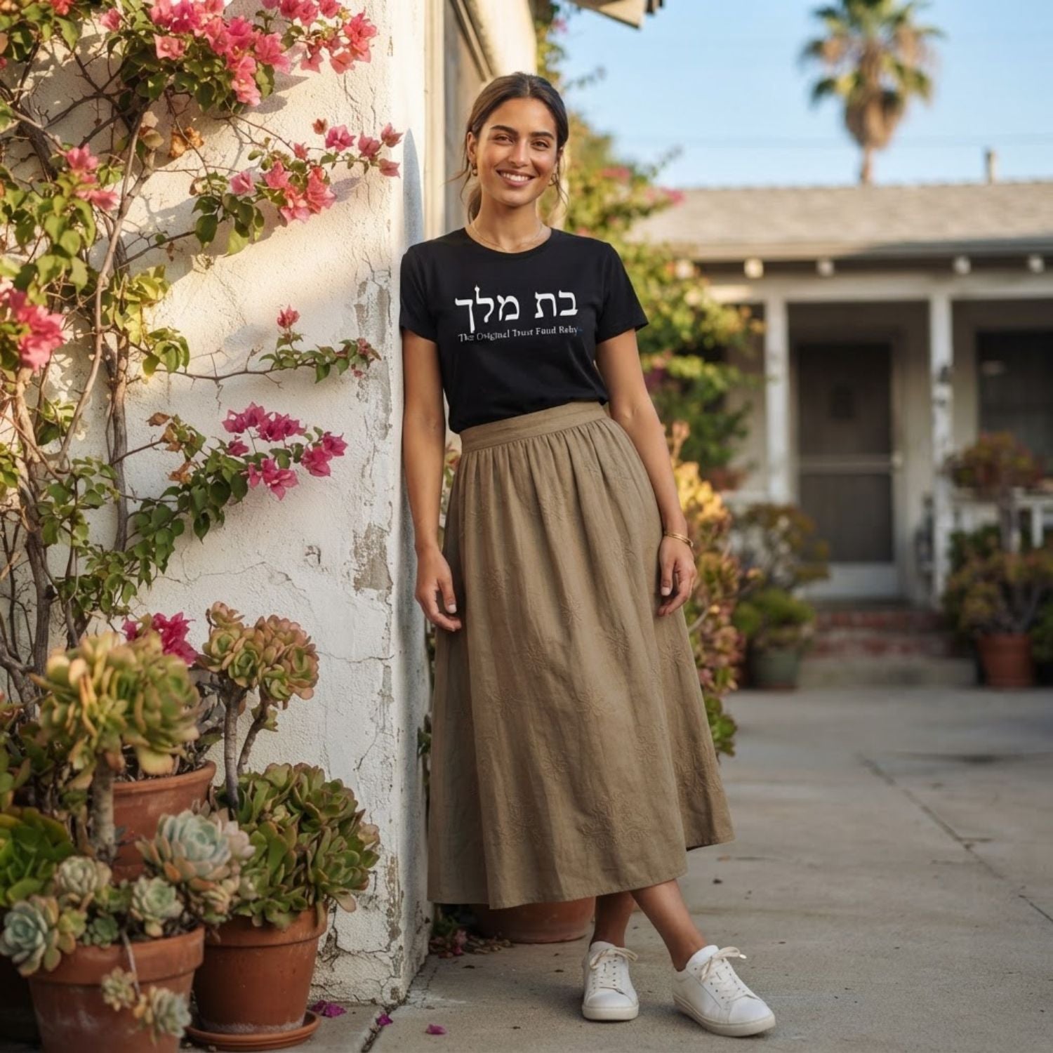 Woman wearing The Original Tee in black by Hashem Is My Sugar Daddy, featuring בת מלך and The Original Trust Fund Baby graphic styled with a long neutral skirt and white sneakers.