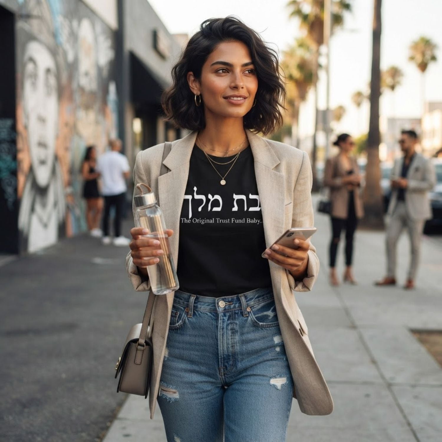 Woman wearing The Original Tee in black by Hashem Is My Sugar Daddy, featuring בת מלך and The Original Trust Fund Baby graphic styled with a beige blazer and blue jeans.