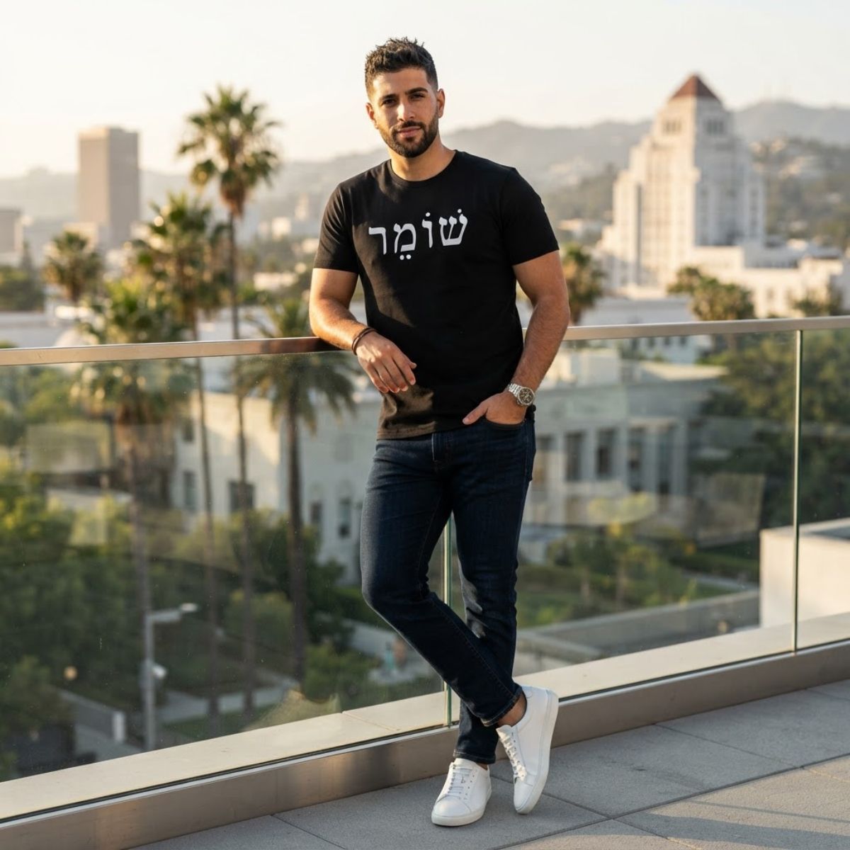 Male model wearing The Protector Tee in black by Hashem Is My Sugar Daddy, featuring שומר Shomer Hebrew protector lettering across the chest, styled with dark jeans on a city rooftop.