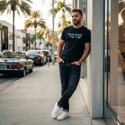Male model wearing The Protector Tee in black by Hashem Is My Sugar Daddy, featuring שומר Shomer Hebrew protector lettering across the chest, styled with dark jeans and white sneakers in a modern street setting.