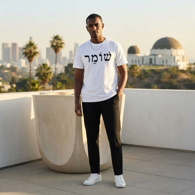 Male model wearing The Protector Tee in white by Hashem Is My Sugar Daddy, featuring black שומר Shomer Hebrew protector lettering across the chest, styled with black pants on a rooftop.