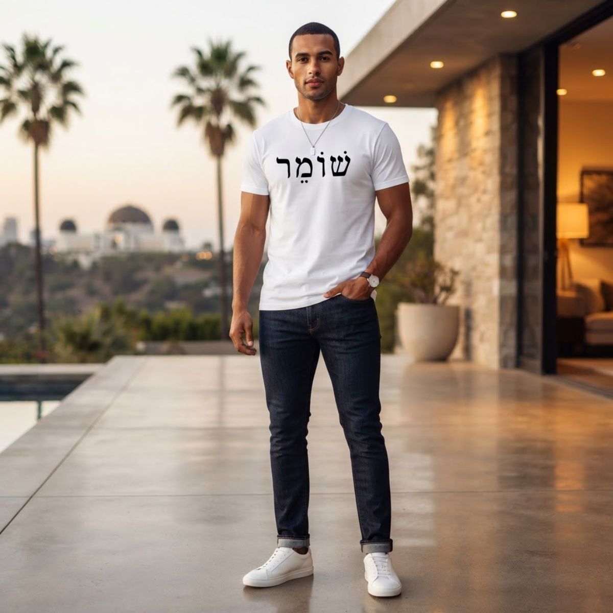 Male model wearing The Protector Tee in white by Hashem Is My Sugar Daddy, featuring black שומר Shomer Hebrew protector lettering across the chest, styled with dark jeans and white sneakers in a modern outdoor setting.