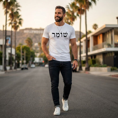 Male model wearing The Protector Tee in white by Hashem Is My Sugar Daddy, featuring black שומר Shomer Hebrew protector lettering across the chest, styled with dark jeans and white sneakers in a modern palm-lined city street setting.